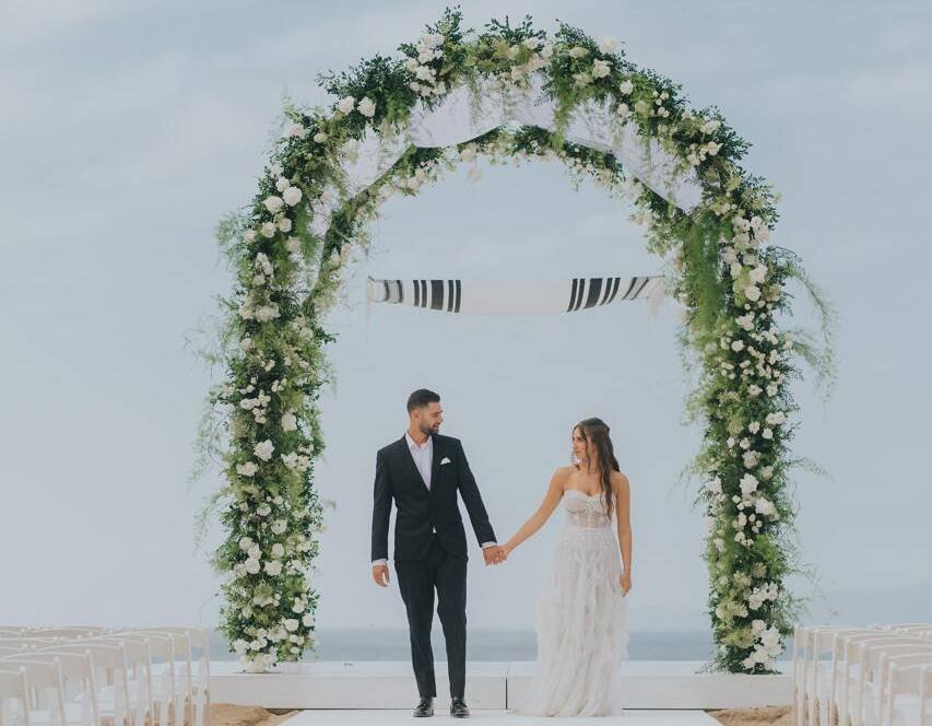 Bride and groom under a floral chuppah with a tallit canopy by the ocean, Jewish destination wedding in Mexico